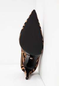 Chaussure à talon stiletto à bout pointu avec semelle noire et dessus rayé tigre, positionnée debout contre un mur blanc d'angle.
