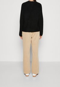 Personne debout de dos, portant un pull à manches longues noir, un pantalon beige à jambes larges et des chaussures blanches sur un fond uni.