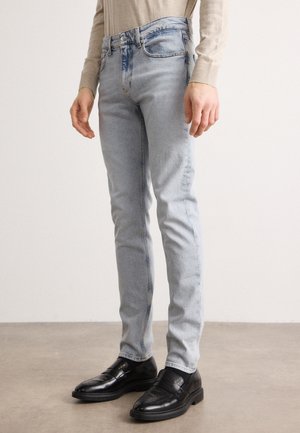 Homme portant un jean slim bleu clair et des mocassins en cuir noir, debout sur un sol en béton lisse devant un mur uni.