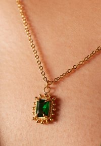 Gouden ketting met een smaragdgeslepen groene steen, omringd door een gedecoreerde gouden setting, ligt op een huidtint achtergrond.