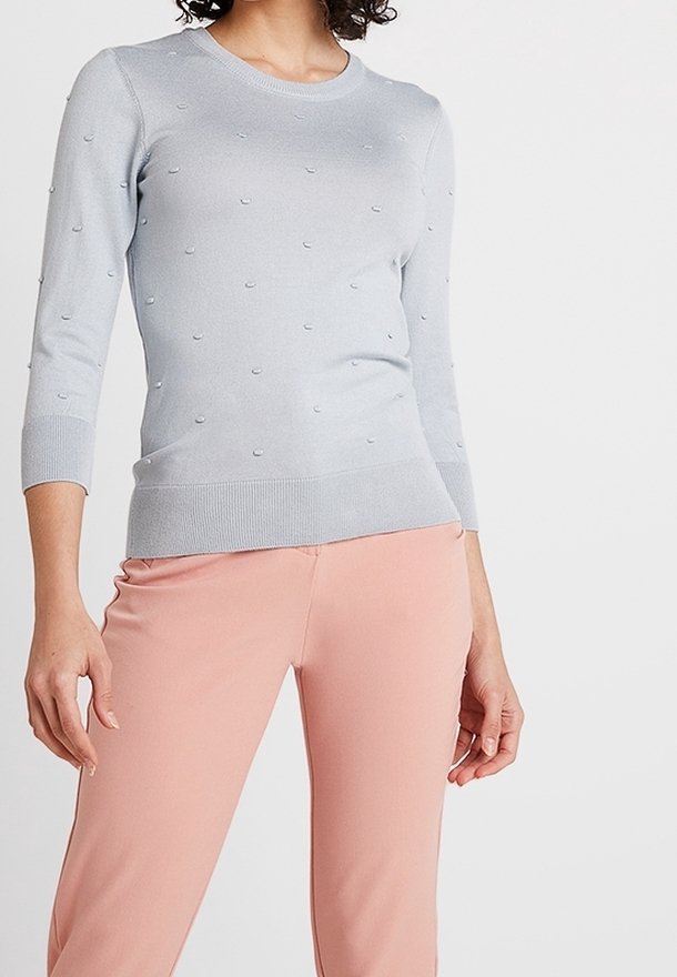 Kvinna som bär en ljusgrå tröja med prickig textur och korta ärmar, matchad med högmidjade, blushfärgade byxor.