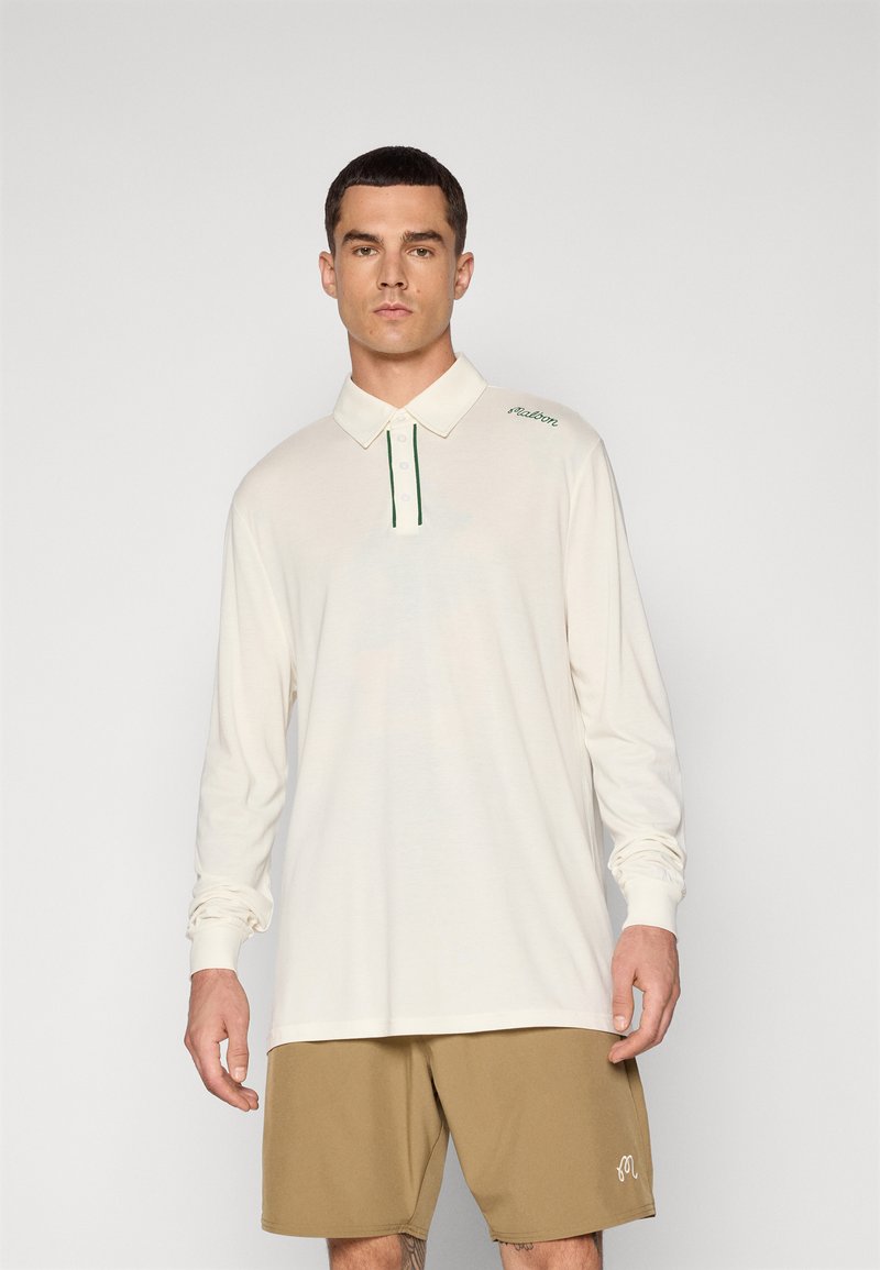 Langermet kremfarget polo skjorte med grønne detaljer og knapper, med krage og brodert logo, kombinert med lysebrune shorts.