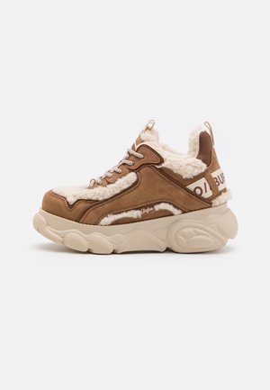 Sneaker marrone in suede con rivestimento bianco soffice, suola chunky beige e lacci metallici. Presenta un logo sul tallone e una parte superiore testurizzata.