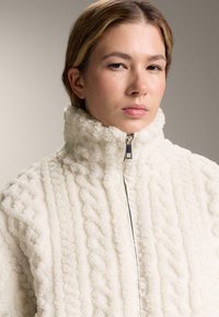 Bílá fleecová bunda s vysokým límcem a strukturovaným vzorem. Disponuje předním zipem a má měkký, útulný vzhled.