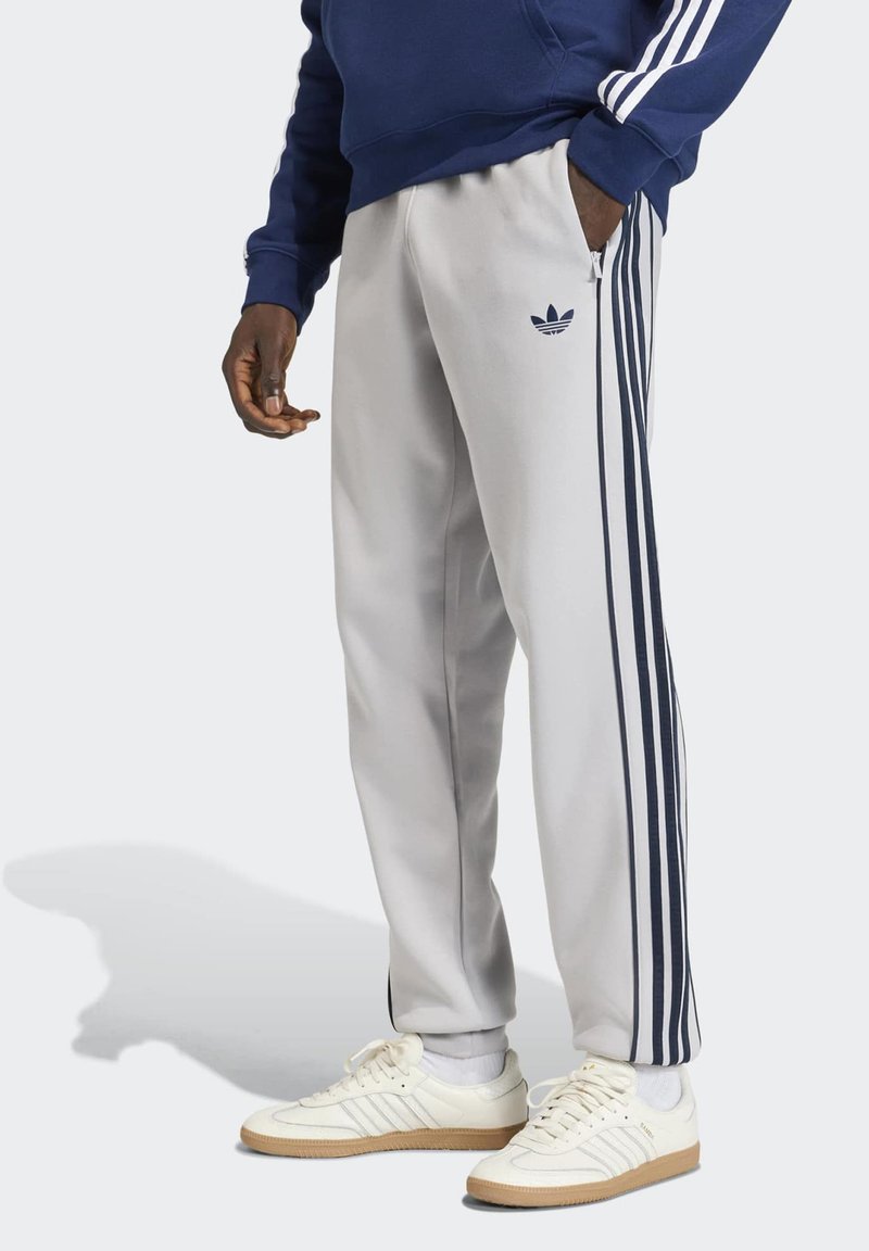 Personne portant un pantalon de jogging Adidas blanc avec des bandes et logos bleu marine, un sweat bleu marine et des baskets Adidas blanches avec semelles en gomme.