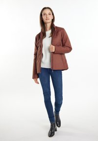 Jachetă insulated de culoare maronie, cu fermoar, guler înalt și buzunare laterale, combinată cu un pulover ușor și blugi albaștri, purtată cu cizme negre la glezne.