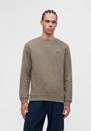 Joven con trenzas, vestido con un sudadera verde oliva lisa y pantalones cargo azul oscuro sobre un fondo blanco.