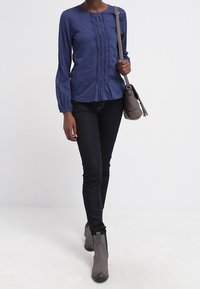 Kvinna som går iklädd en blå långärmad blus, svarta tajta jeans, grå ankelstövlar och bär en grå axelväska.