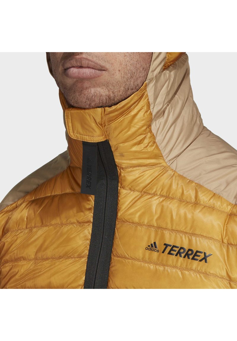 Gelbe und beige isolierte Jacke mit hohem Kragen, gestepptem Design und schwarzen Hardware-Akzenten, mit dem "Terrex"-Logo.