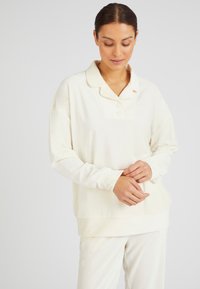 Sudadera de gran tamaño en color crema con cuello tipo polo, cierre de tres botones y textura acanalada. Presenta mangas largas y un ajuste holgado.