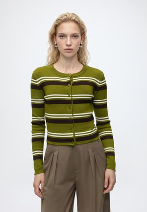 Femme blonde portant un cardigan boutonné rayé vert, noir et blanc et un pantalon plissé marron, debout devant un fond blanc uni.