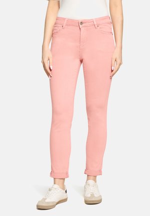 Kobieta w jasnoróżowych, obcisłych spodniach z podwiniętymi nogawkami i białych trampkach, stojąca na tle jednolitego, jasnego tła.