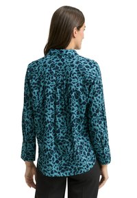 Türkisfarbene Bluse mit schwarzen Leopardenflecken, langen Ärmeln und Kragen. Am Rücken mit einer Falte versehen. Glatte Textur.