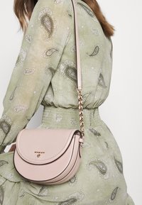 Růžová kožená crossbody taška s zakřiveným klopovým víkem, zlatým kováním a řetízkem. Pozadí tvoří zelené šaty s paisley vzorem.