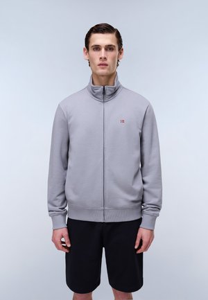 Ung mand står fremadrettet iført grå lynlåstrøje med lille flaglogo og sorte shorts mod en ensfarvet hvid baggrund.