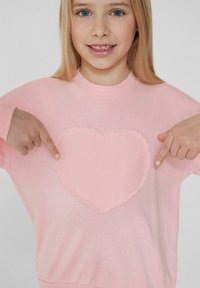 Světle růžová mikina s velkým srdcovým nášivkem na přední straně s roztrhanými okraji. Design má kulatý výstřih a široké rukávy.