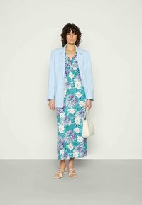 Blazer azul sobre um vestido maxi de estampado floral com flores turquesa, brancas e roxas. Modelo carrega uma bolsa branca, usando sandálias com tiras.