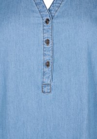 Hele sinine denim-särk, millel on V-kaelus, kolm nuppu ja nähtavad õmblused krae ja nööbirea ääres. Sile tekstuur.