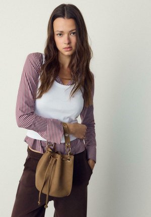 Jeune femme en chemise rayée et débardeur blanc tenant un sac seau en daim marron avec des anneaux métalliques, portant un pantalon foncé sur un fond uni.