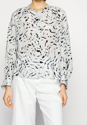 Personne portant un chemisier à manches longues et col rond avec des motifs abstraits ondulés noirs et gris et un pantalon blanc sur un fond uni.