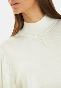 Lichtbeige trui met een hoge ribbelkraag, voorzien van gathered stof en kleine parelaccenten bij de halslijn. Glad textuur.