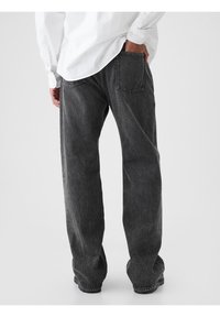 Jean en denim noir coupe décontractée avec une finition légèrement délavée, des coutures visibles et deux poches arrière, porté avec une chemise blanche à manches longues.