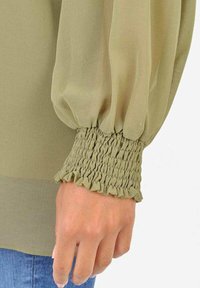 Blusa verde oliva a maniche lunghe con maniche a sbuffo e polsini arricciati. Tessuto morbido con un tocco di ruffles al polso.