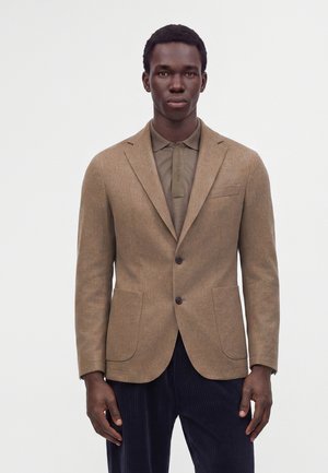 Homme portant un blazer beige sur un polo marron et un pantalon foncé, debout devant un fond clair uni.