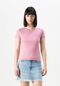 Femeie cu păr drept și închis la culoare, purtând un top roz ajustat cu mânecă scurtă și o fustă din denim deschis la culoare, ușor uzată, pe un fundal simplu.