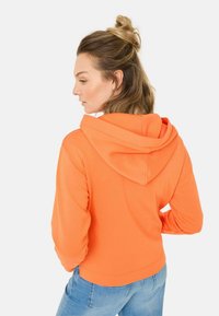 Felle oranje cropped hoodie met een ontspannen pasvorm, verstelbare trekkoordkap, en zachte textuur, gecombineerd met lichtblauwe jeans.