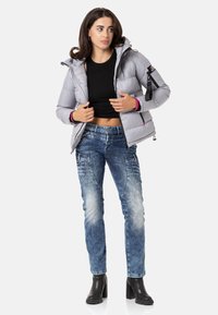 Hellgraue Pufferjacke mit Kapuze, ausgestattet mit schwarzen Reißverschlüssen und rosa Akzenten an den Ärmeln. Kombiniert mit blauen, verwaschenen Jeans und schwarzen Stiefeletten mit Absatz.