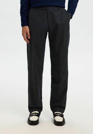 Homme portant un pantalon droit gris foncé, des mocassins noir et blanc, et un pull bleu marine, debout les mains dans les poches.