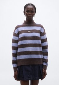 Barna és világoskék csíkos kötött pulóver magas nyakkal, amelyen egy kis logó folt látható a mellkasnál, és bordázott mandzsetták. Farmer szoknyával párosítva.