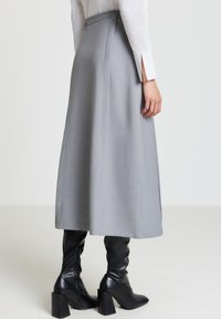 Grauer Midirock aus glattem Stoff mit tailliertem Schnitt. Mit Bund und dezenten Schlitzen an den Seiten; kombiniert mit kniehohen schwarzen Stiefeln.