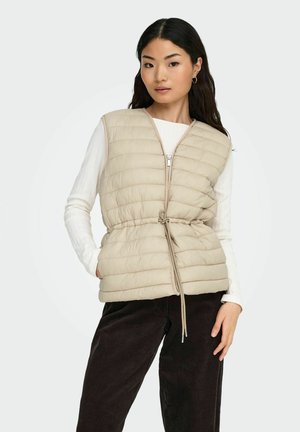 Jauna moteris dėvi kreminės spalvos siuvinėtą liemenę be rankovių ant baltos ilgarankovės palaidinės ir tamsiai rudų kelnių, stovinti viena ranka kišenėje.