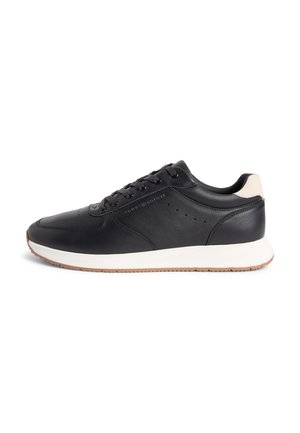 Zwarte lage leren sneaker met witte zool, beige hieltab, zwarte veters en "Tommy Hilfiger" branding aan de zijkant.