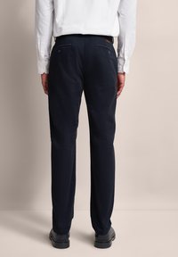 Mężczyzna w czarnych spodniach, czarnych butach i białej koszuli z długim rękawem, pokazany z tyłu na jednolitym tle.