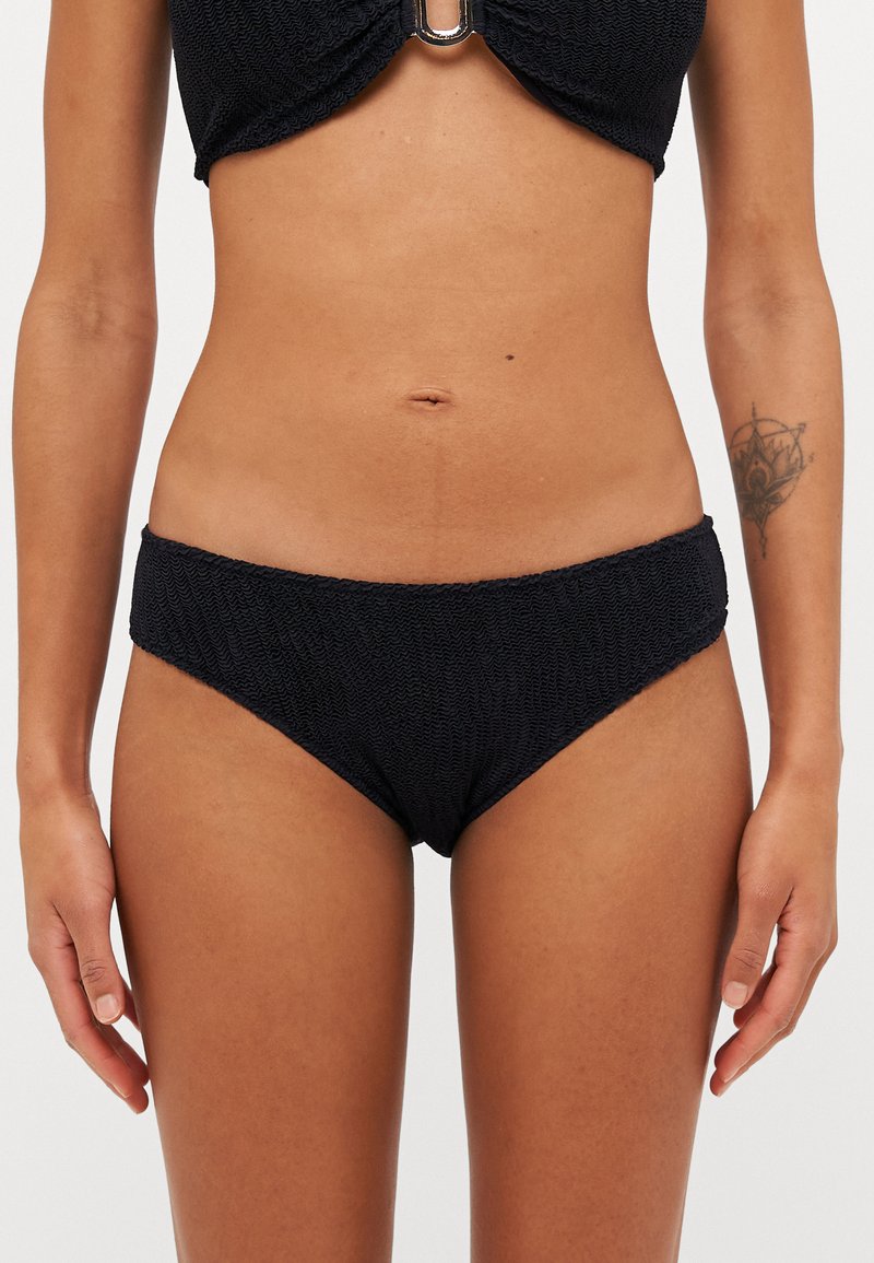 Melna bikini komplekts ar teksturētu augšdaļu un zemu vidukļa apakšdaļu. Audumam ir aušanas raksts, un augšdaļā ir centrālais metāla akcents.