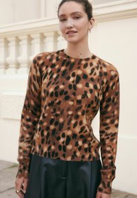 Femme portant un pull en imprimé léopard marron et noir et un pantalon noir, se tenant à l'extérieur près d'une balustrade couleur crème.