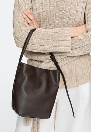 Person iført beige strikket sweater holder en mørkebrun læderbuckettaske med en lang justerbar rem over hvide bukser.