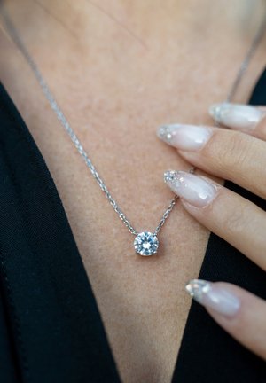 Silberne Kettenhalskette mit einem runden, klaren Edelsteinanhänger. Eine Hand ruht daneben und zeigt lange, manikürte Nägel mit einem glitzernden Überlack.