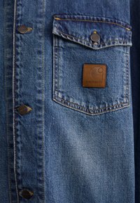 Plava traper košulja s džepom na prsima, metalnim gumbima i smešnim kožnim Carhartt logom. Vidljive šavove i detalji teksture.