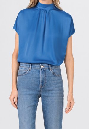 Femme portant un chemisier bleu à manches courtes et à col montant, rentré dans un jean taille haute bleu clair, debout devant un fond uni.