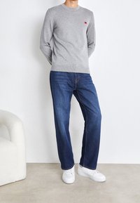 Szary sweter z okrągłym dekoltem i małym czerwonym logo, zestawiony z niebieskimi dżinsami i białymi trampkami, stojący na jasnym tle.