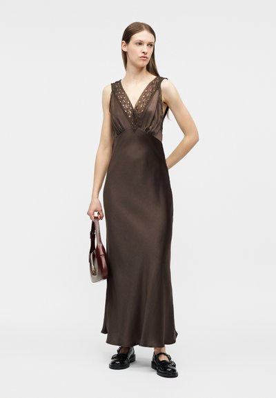 Γυναίκα φορώντας ένα αμάνικο σκούρο καφέ σατέν φόρεμα με δαντέλα, κρατώντας μια μικρή καφέ τσάντα και φορώντας μαύρα παπούτσια.