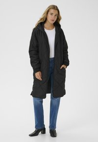 Langer schwarzer steppschirmmantel mit Kapuze, Frontreißverschluss und seitlichen Taschen, kombiniert mit blauen Jeans und schwarzen Stiefeletten. Glatte Textur.