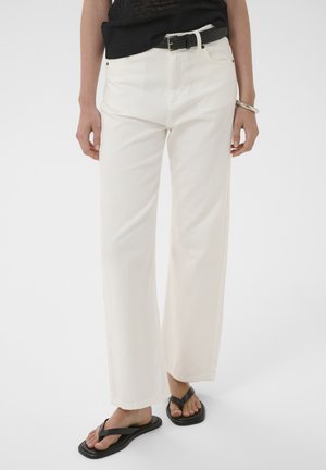 Nő, aki törtfehér, magasderekú egyenes szárú nadrágot, fekete övet, fekete felsőt, ezüst karkötőt és fekete flip-flop papucsot visel fehér háttéren.