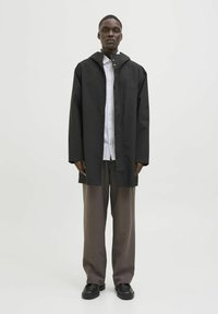 Oferind o protecție împotriva ploii, acest trench ușor de culoare neagră, cu glugă și închidere cu capse, este purtat peste o cămașă albă cu nasturi și pantaloni gri. O pereche de pantofi negri completează ținuta.