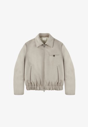 Chaqueta beige con cremallera y textura suave, presenta un cuello con solapa, dobladillo elástico, bolsillos laterales y un pequeño bolsillo en el pecho con un botón.