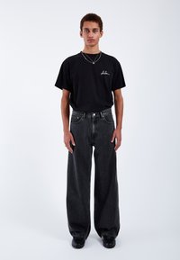 Černé oversized tričko s bílým vyšívaným logem, spárované s volně střiženými černými džíny, nošeno s černými botami na neutrálním pozadí.